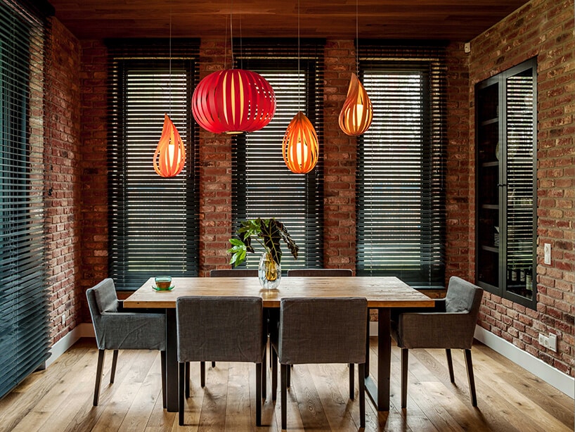 mała jadalnia z ceglanymi ścianami z wysokimi wąskim oknami z czarnymi drewnianymi żaluzjami wewnętrznymi z drewnianym stołem pod czerwonymi lampami