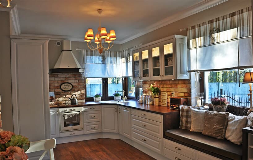kuchnia w stylu prowansalskim z białymi frontami ścianami z ceglanymi wstawkami z ciemnym drewnianym blatem z siedziskiem przy dużym oknie na końcu blatu kuchennego