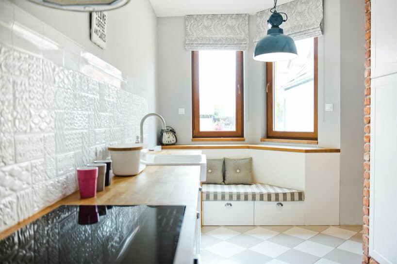 nowoczesna biała kuchnia z fakturowanymi białymi płytkami nad drewnianym blatem kuchennym na tle siedziska pod dwoma wąskimi oknami