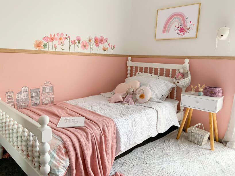 duże białe łóżko i mały stolik na tle ózowej ściany i namalowanymi na niej kwiatkami