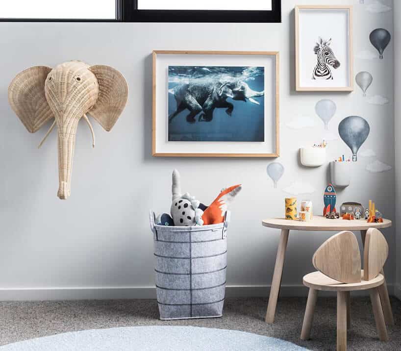 pokój dziecięcy z drewnianym stolikiem i krzesłem na tle ściany z namalowanymi balonami i obrazkami w ramkach i wiklinową głową słonia