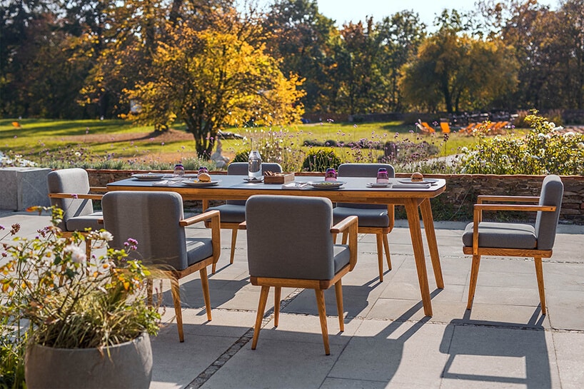 elegancki drewniany stół z szarym blatem z sześcioma drewnianym krzesłami z szarym siedziskiem i oparciem na tarasie na tle rozległego ogrodu