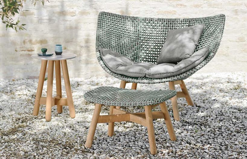 elegancki zestaw Mbrace od Dedon fotel i podnóżek z drewnianą kostrukcją i szarym plecionym siedziskiem obok małego drewnianego stolika